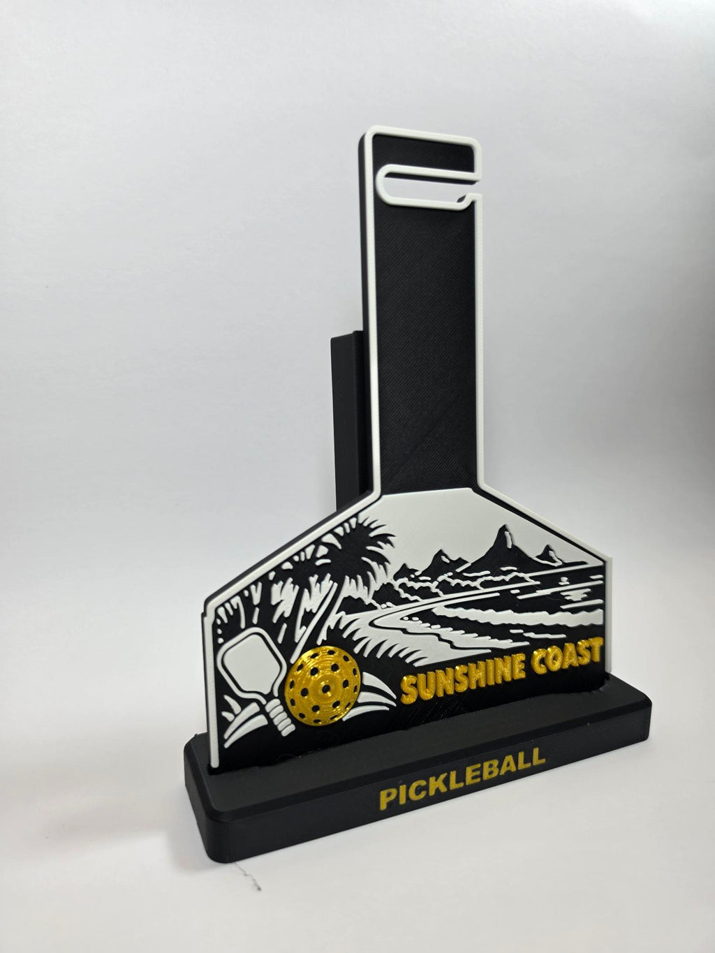 A pickleball-themed decorative plaque or stand with a black base and backboard-shaped front panel. The front features a white illustrated beach scene with palm trees, mountains, a paddle, a yellow pickleball, and the words 'SUNSHINE COAST' and 'PICKLEBALL' in yellow.